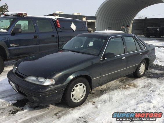 Dark sedan parked next to a blue pickup truck in a snowy lot. A large arched structure is in the background
