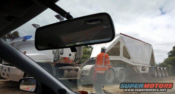 View from inside a car looking at two large trucks, one with a white trailer, and a worker in an orange jacket