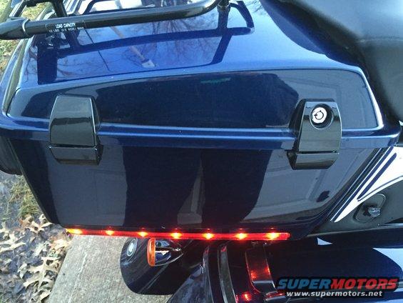 Close-up of a blue motorcycle saddlebag with black latches and a red LED light strip underneath. Text on a rack above reads 'LOAD CAPACITY'