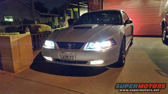 Front view of a silver 2002 Ford Mustang parked in a driveway at night. Headlights are on