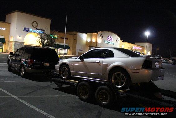 White 2002 Ford Mustang coupe on a trailer, towed by a black SUV. Post Office and discount store in background