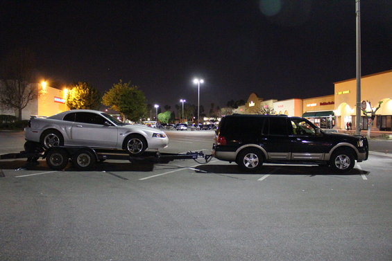 Dark blue Ford Expedition towing a silver Ford Mustang on a trailer at night in a parking lot