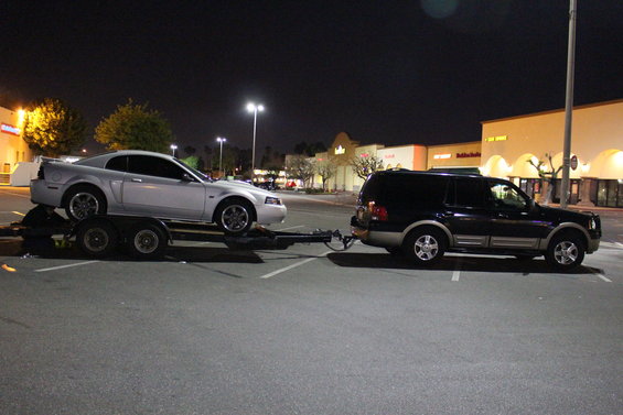 Silver 2003 Ford Mustang on trailer towed by black Ford Expedition at night