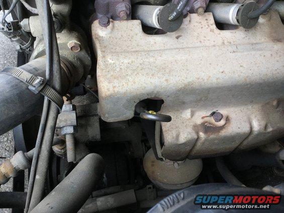 Close-up of engine parts, including hoses, metal covers, and spark plug wires. A yellow ring is visible on one component