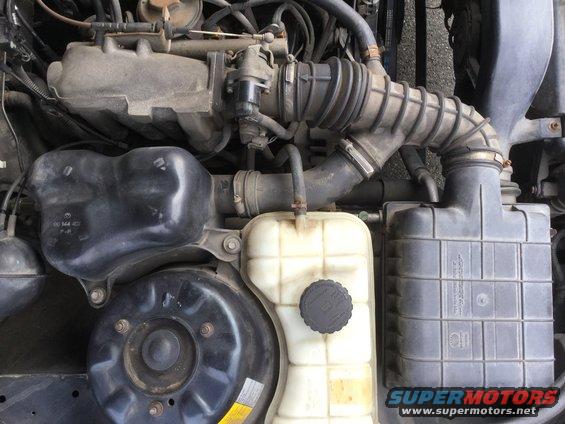 Engine bay with air intake, coolant reservoir, and other parts visible. Some text is present on components