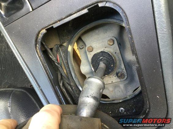 Gear shifter mechanism inside a car interior, showing linkage and wiring. A hand is visible holding the shifter