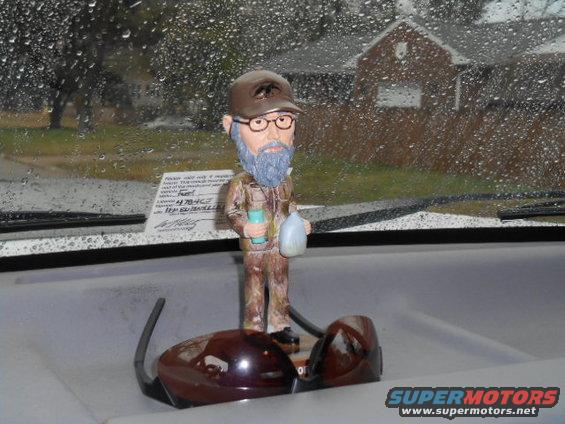 Bobblehead figure with glasses and a hat sits on a dashboard in front of a rain-streaked windshield