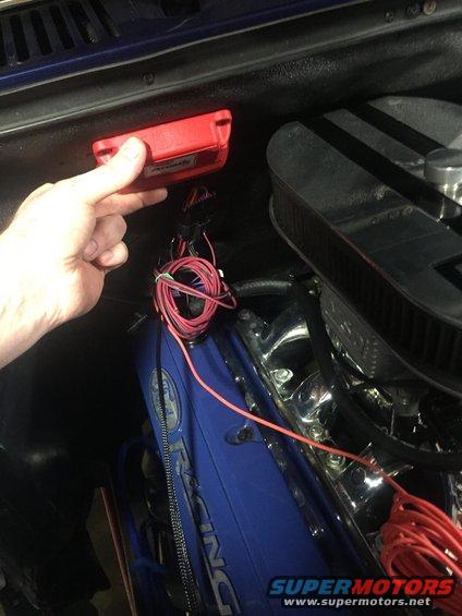 Hand holding red electronic module near engine wiring and blue valve cover with 'Racing' text