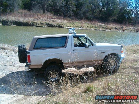 1996 Ford Bronco with aftermarket lights on roof, parked on rocky terrain near a river