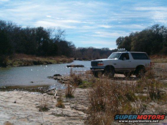 White Ford Bronco parked by a river with brush and trees in the background