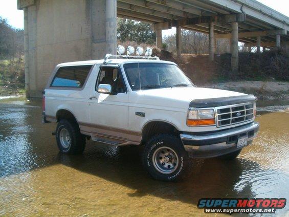 White Ford Bronco with aftermarket lights on roof rack, parked in shallow water under a bridge