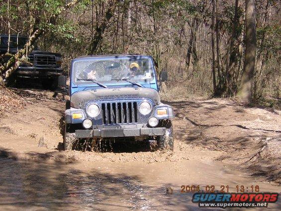 Blue Jeep Wrangler driving through mud puddle, another vehicle behind