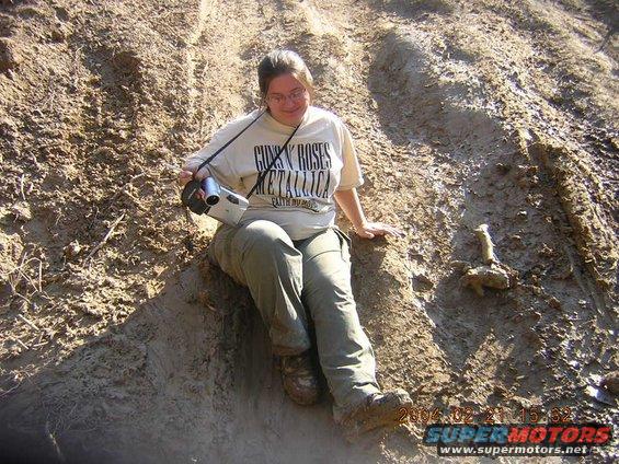 Person in Guns N' Roses t-shirt and cargo pants sits on a muddy hill, holding a camcorder. Date stamp 2004-02-21