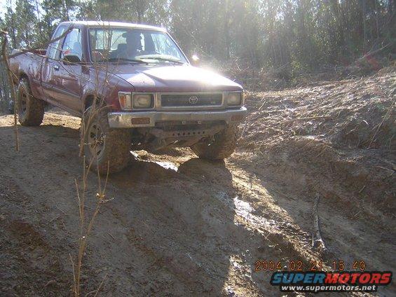 Red pickup truck on a muddy incline, front 3/4 view. The vehicle has off-road tires and appears to be stuck