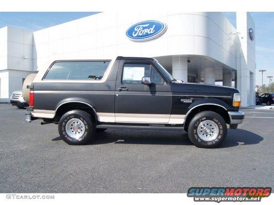 Side view of a dark gray Ford Bronco with a tan top and trim. It's parked in front of a Ford dealership