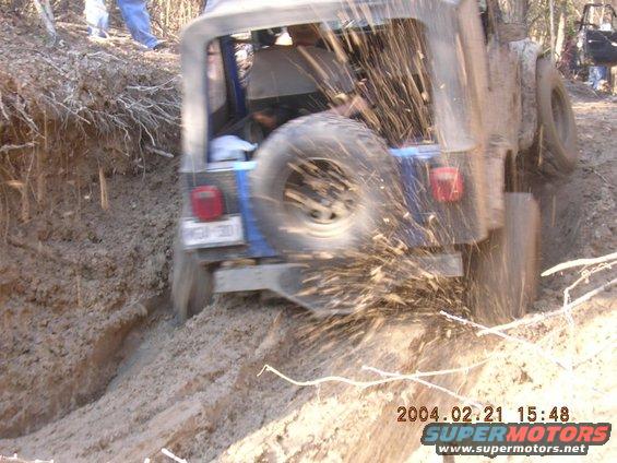 Blue Jeep Wrangler driving through mud, rear view. Another vehicle visible in background