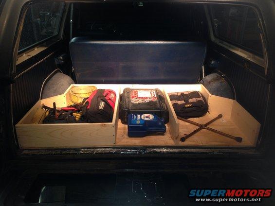 Custom wooden storage box with tools, bags, and a jack in the cargo area of an older SUV