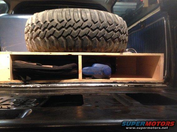 Spare tire on wooden storage box inside a vehicle cargo area. A blue tool is in the box