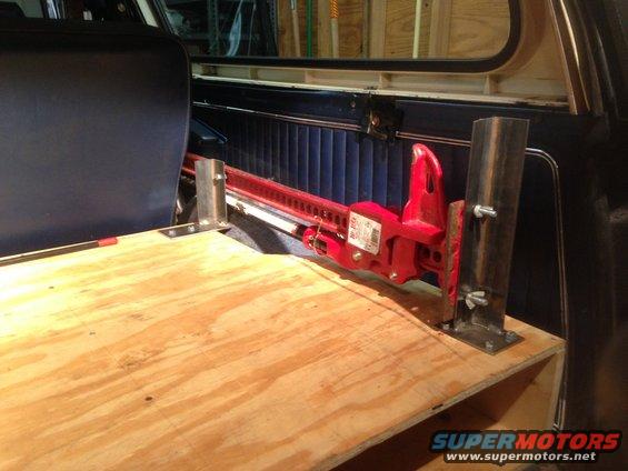 Red hi-lift jack mounted to custom wooden platform inside vehicle, next to dark blue seat