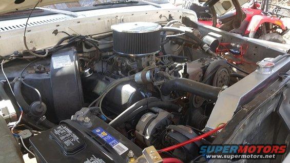 Engine bay with V8 engine, air filter, battery, and radiator visible. Some red ATV in background