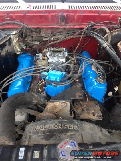 Engine bay with blue valve covers and a carburetor. Radiator fan shroud has a caution label
