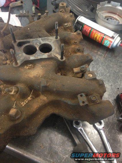 Dirty engine intake manifold with two carburetor mounting holes and several bolts. A can of brake cleaner and wrenches are nearby