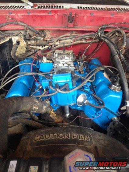 Engine bay with blue painted intake manifold and valve covers, carburetor, and fan shroud with caution text