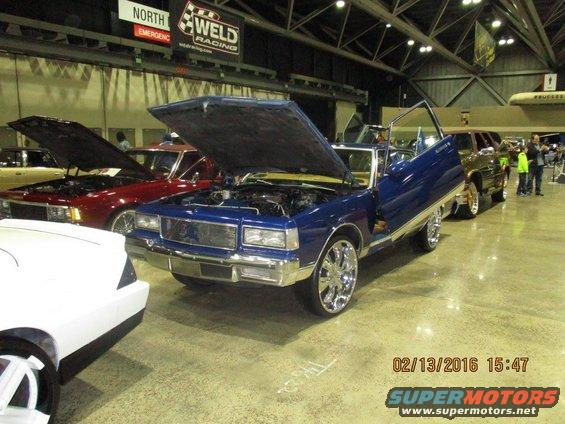 Blue sedan with gullwing doors and chrome rims, hood open, parked indoors next to other cars