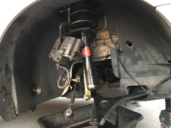 Close-up of car's front suspension with brake caliper, strut, and tie rod end visible. Oil drain pan underneath