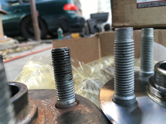 Two wheel studs, one rusty and one clean, in the foreground with a green car in the background