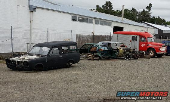 Several derelict vehicles, including a black van, a green car body, and a red truck, sit in an outdoor lot