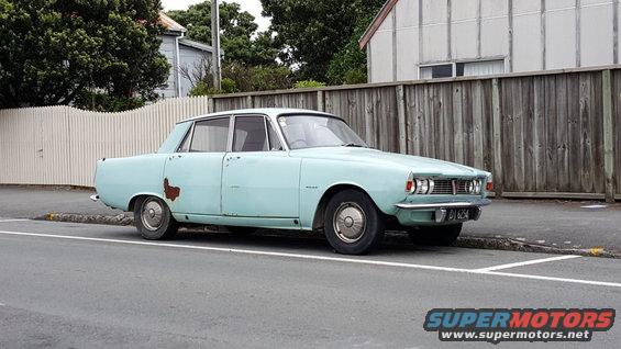 light blue sedan with rust spots on the side, parked on a street next to a wooden fence