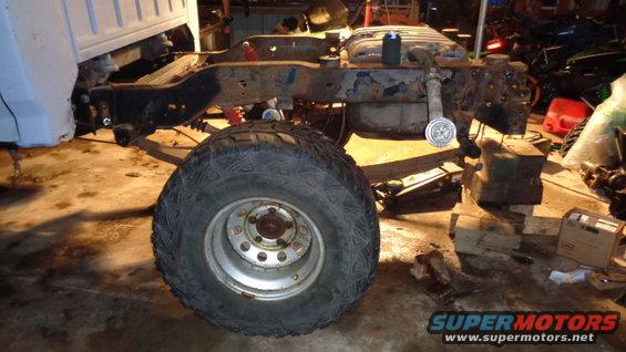 Truck frame with suspension components and a large tire, resting on blocks in a garage