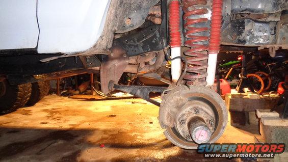Close-up of a vehicle's front suspension and brake components, with red shocks and coil springs visible. The wheel hub is exposed
