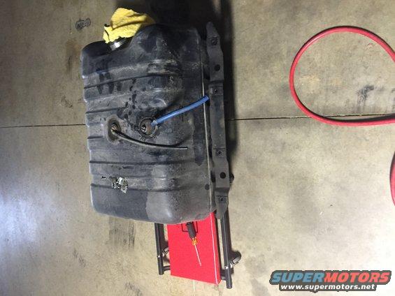 Vehicle fuel tank with blue hose and yellow rag on top, mounted on a bracket. Red toolbox below