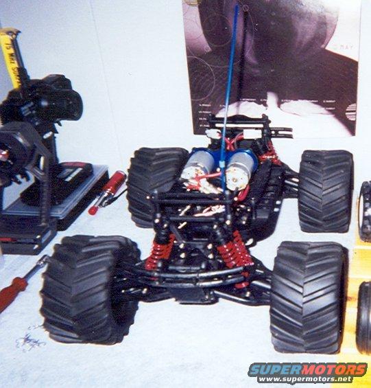 Radio-controlled monster truck chassis with dual motors, red shocks, and large tires. Remote control transmitter is visible to the left
