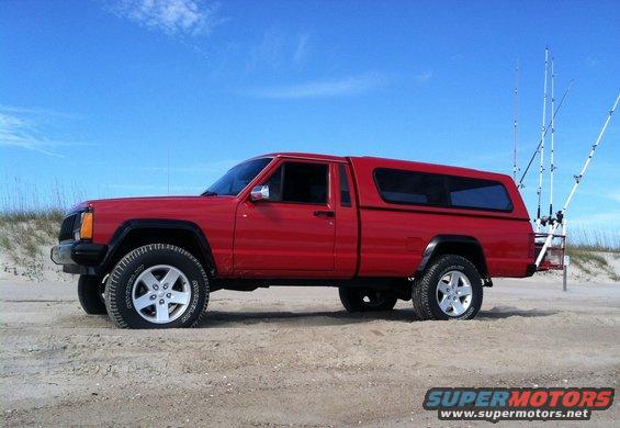 red Jeep Comanche pickup truck with camper shell parked on a sandy beach with fishing rods in the back