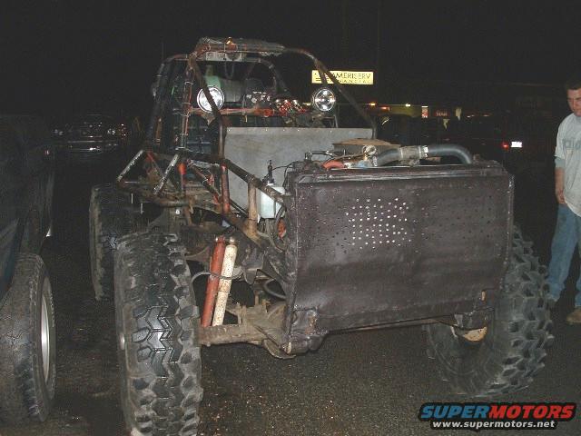 Custom off-road buggy with large tires, exposed engine, and roll cage, parked next to another vehicle at night