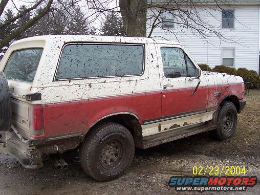 Mud-splattered red and white Ford Bronco parked outside a house. Spare tire mounted on rear