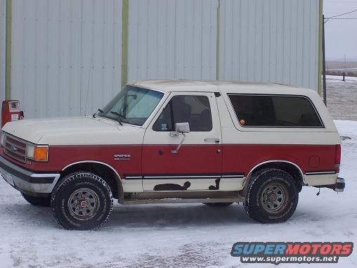 Two-tone red and white 1987 Ford Bronco in snow, side view, showing rust and wear
