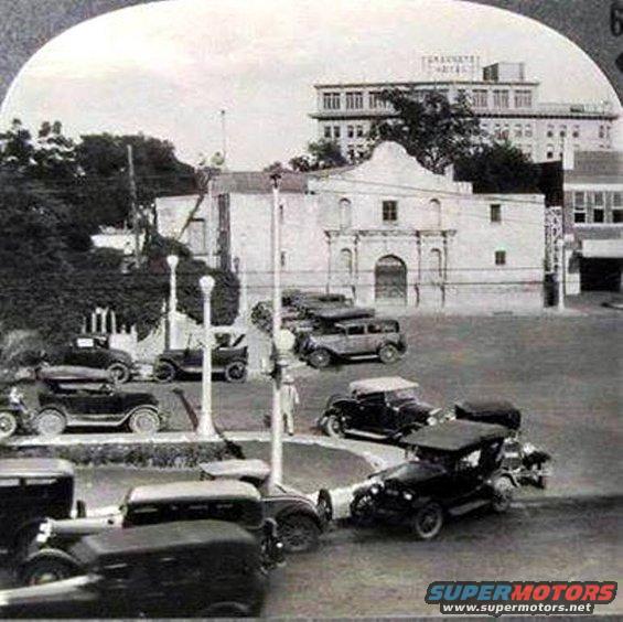Several vintage cars parked in front of the Alamo building with a hotel in the background