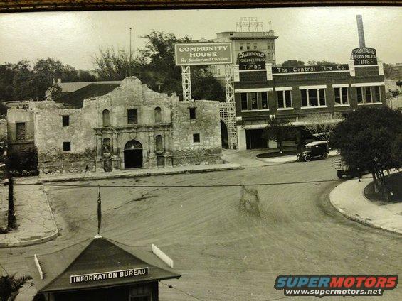 View of the Alamo and buildings, including a tire company and community house, with a couple of cars on the street