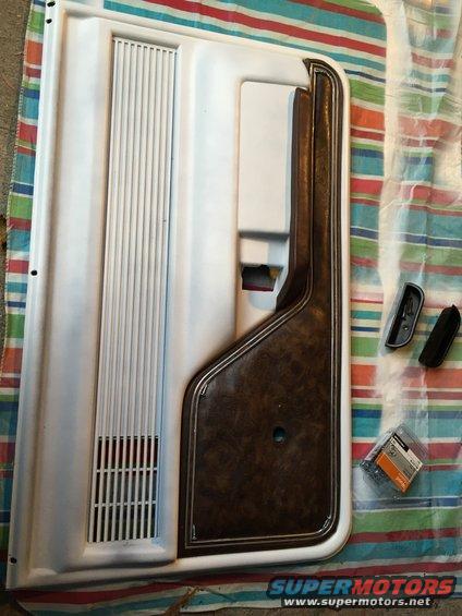 White car door panel with wood grain insert and speaker grille, next to door handles and hardware