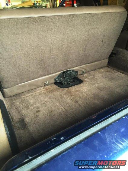 Back seat folded down in a vehicle, showing the carpeted floor and seat base. There's a metal latch mechanism visible