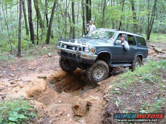 Dark green Toyota 4Runner driving through a muddy ditch in a wooded area. Two people are inside