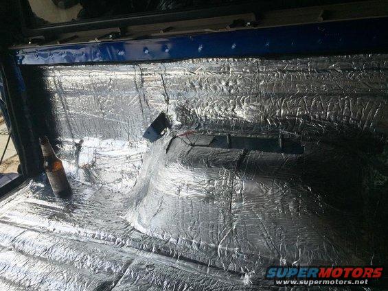 Interior of a vehicle with soundproofing material applied to the floor and rear panels. A bottle of beer is on the floor