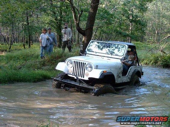 White Jeep CJ driving through a muddy creek with people watching from the bank