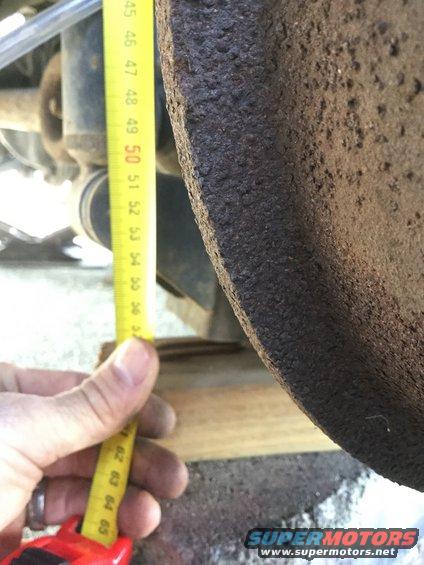Hand holding a yellow measuring tape against a rusty brake rotor. Tape shows markings from 45 to 65