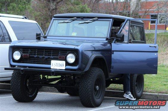 Blue Ford Bronco, driver's door open, parked next to a white SUV. License plate reads 'A STUD69'