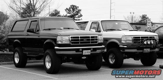Two lifted Ford Broncos parked side-by-side. The one on the left is black, the one on the right is white with a brush guard